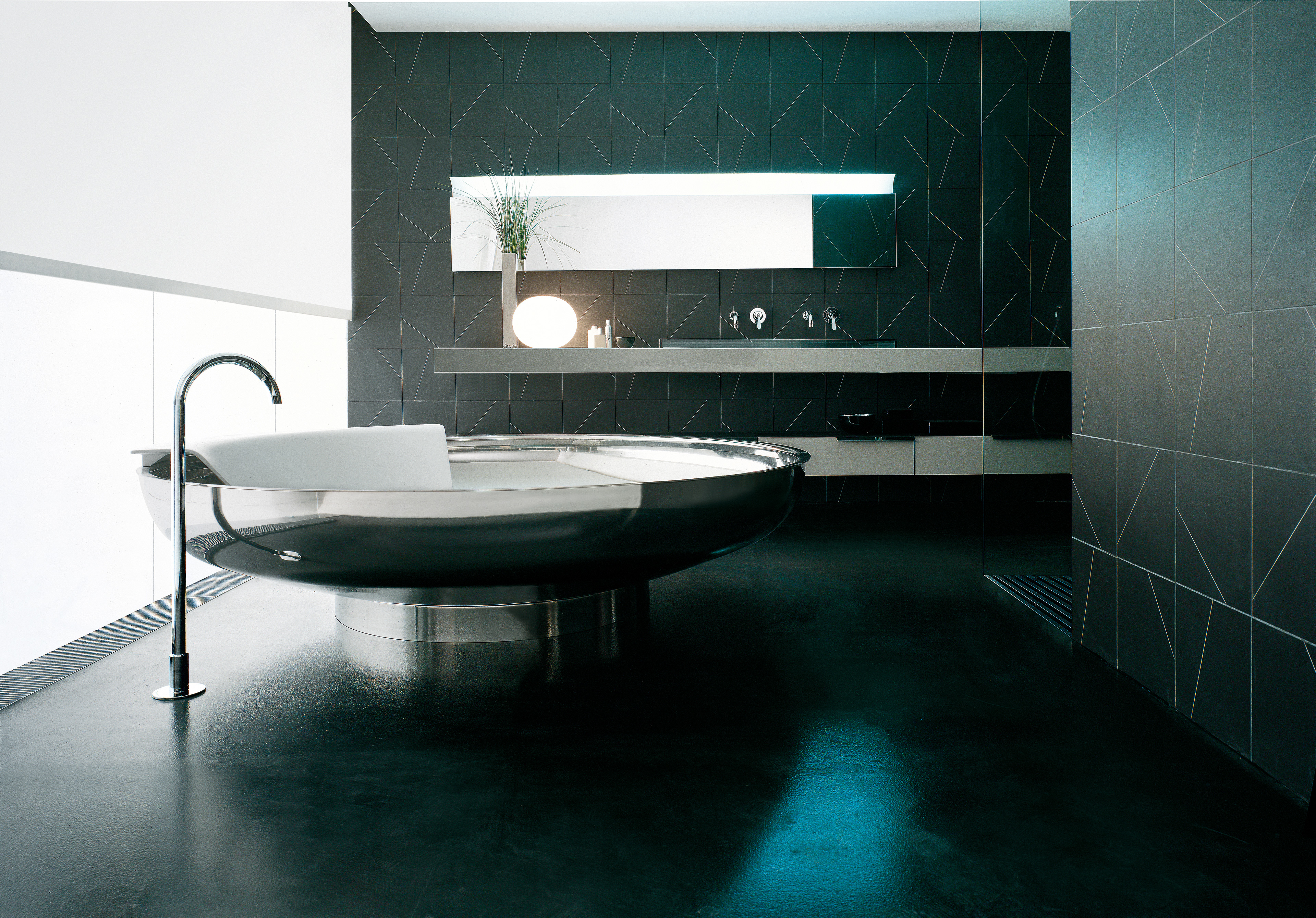 Runde freistehende Badewanne Ufo von Agape in dunkelgrau und weiß, aus glänzendem Edelstahl mit Cristalplant, Design Benedini Associati, in einem modernen Badezimmer mit dunklen Fliesen und Armatur.