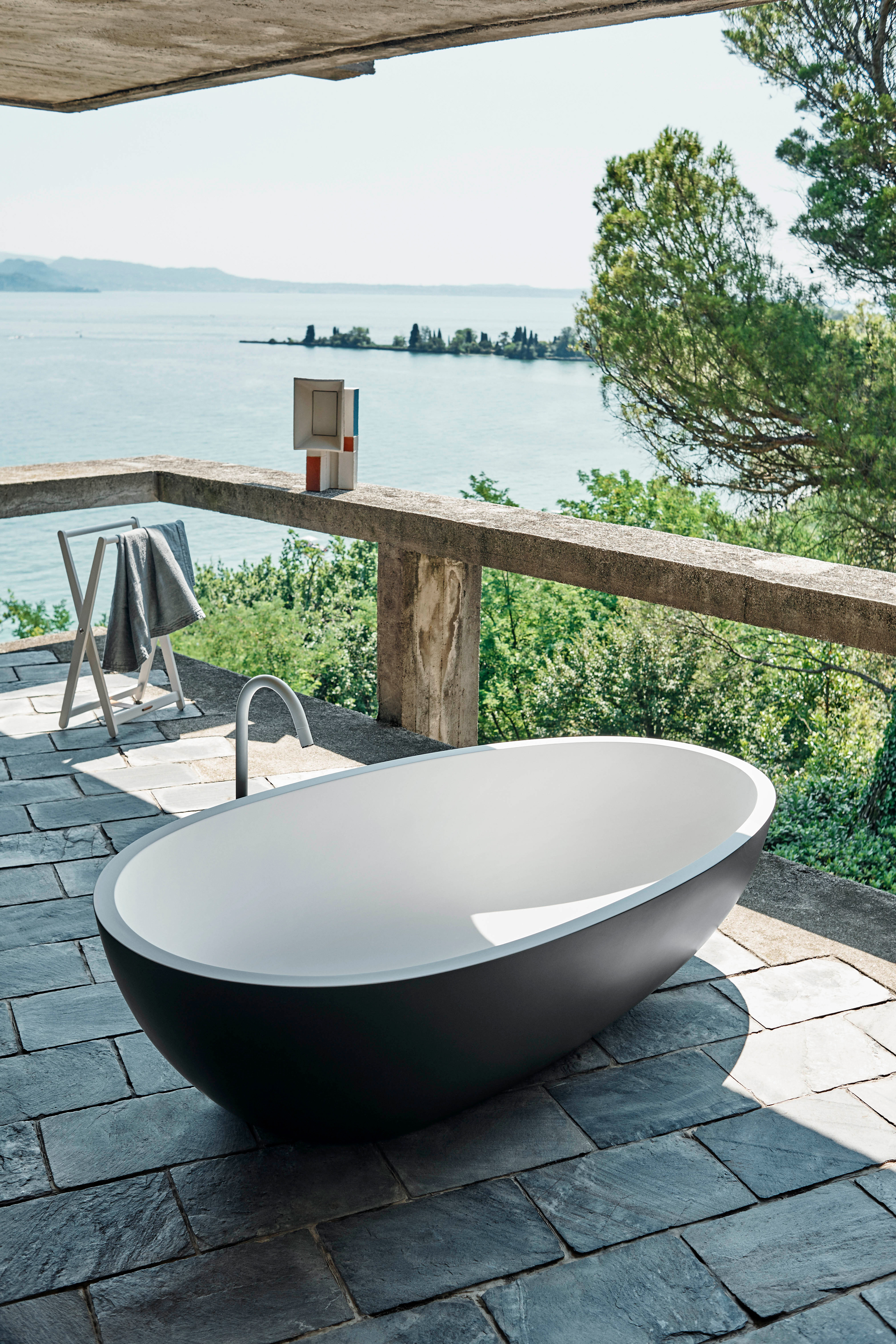 Freistehende Badewanne Spoon M von Agape in dunklem Grau und Weiß, zweifarbig, aus Cristalplant biobased Mineralguss, Design Benedini Associati, platziert auf einem Balkon mit Blick auf ein Gewässer.
