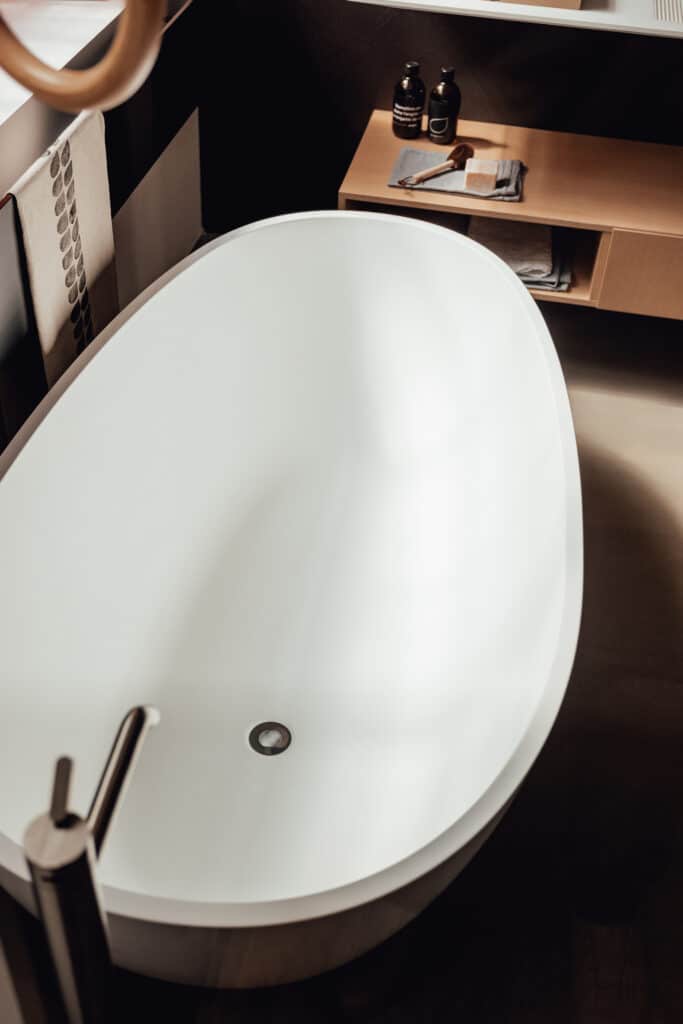 Freistehende Badewanne Spoon M von Agape in reinem Weiß aus Cristalplant biobased Mineralguss, Design Benedini Associati, in einer Aufsicht im Badezimmer mit Armatur in glänzendem Metall und hölzernen Bad-Accessoires.